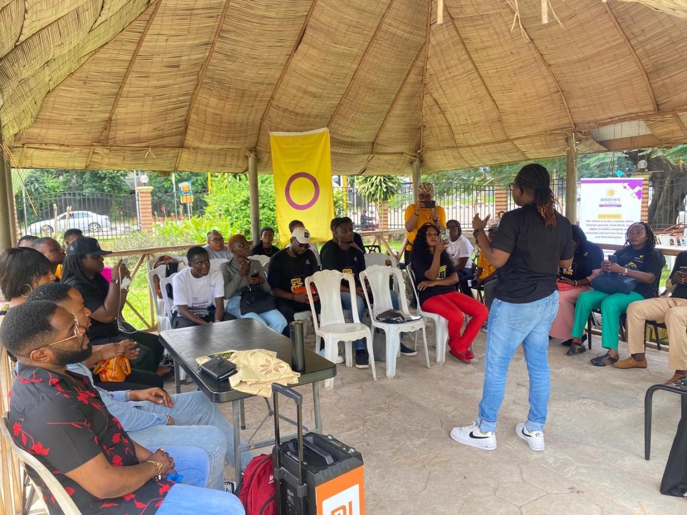 Team members from Intersex Nigeria gather outside in a pavilion, some seated in a semi-circle, with an intersex flag behind them