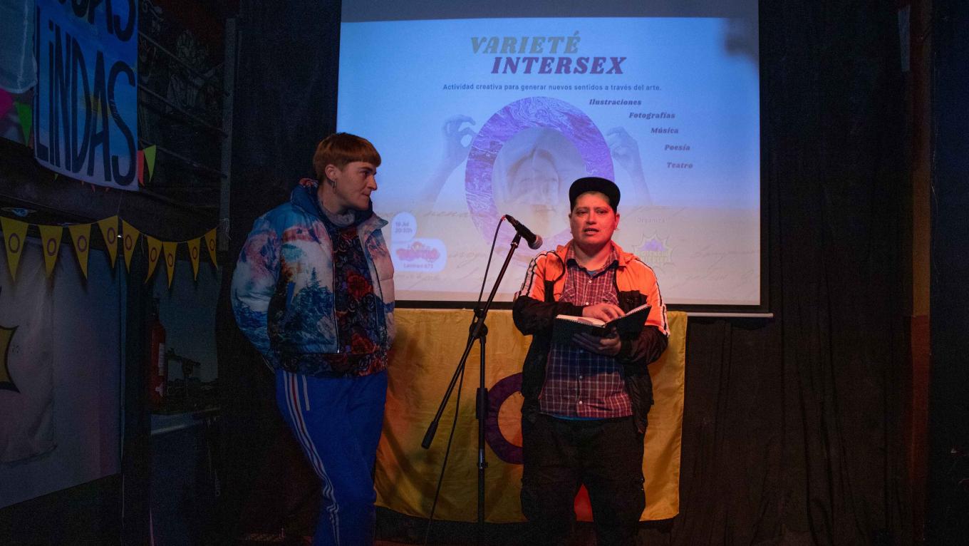 Two speakers stand in front of a projector screen which reads 